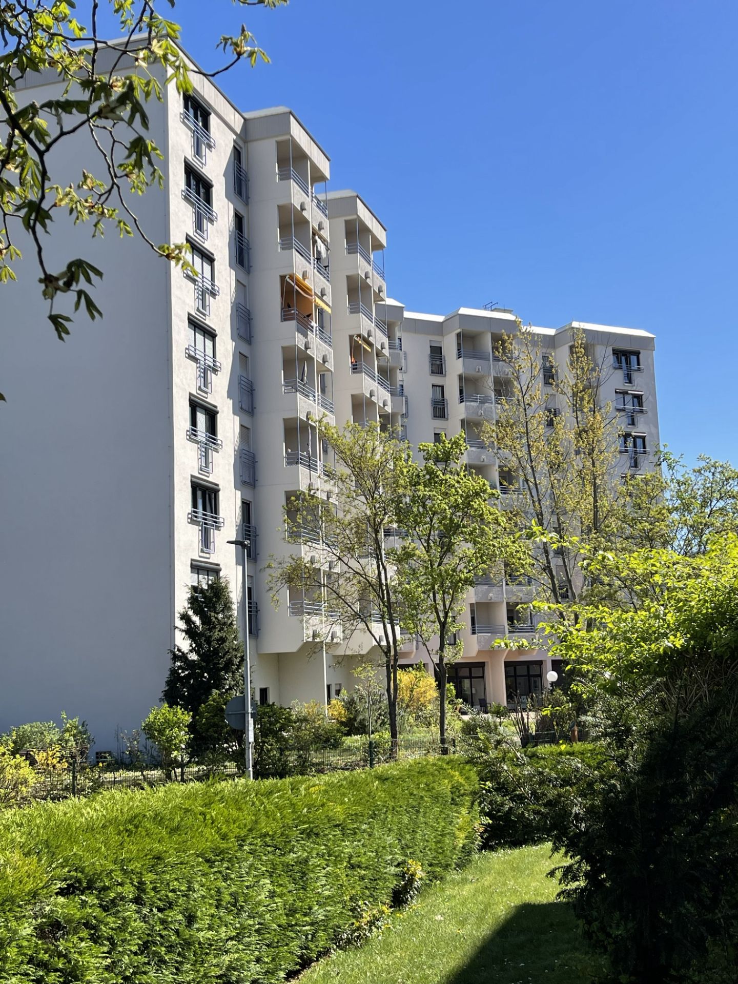 Mehrstöckiges Wohnhaus mit Balkonen, umgeben von Bäumen und Grünanlage bei blauem Himmel.