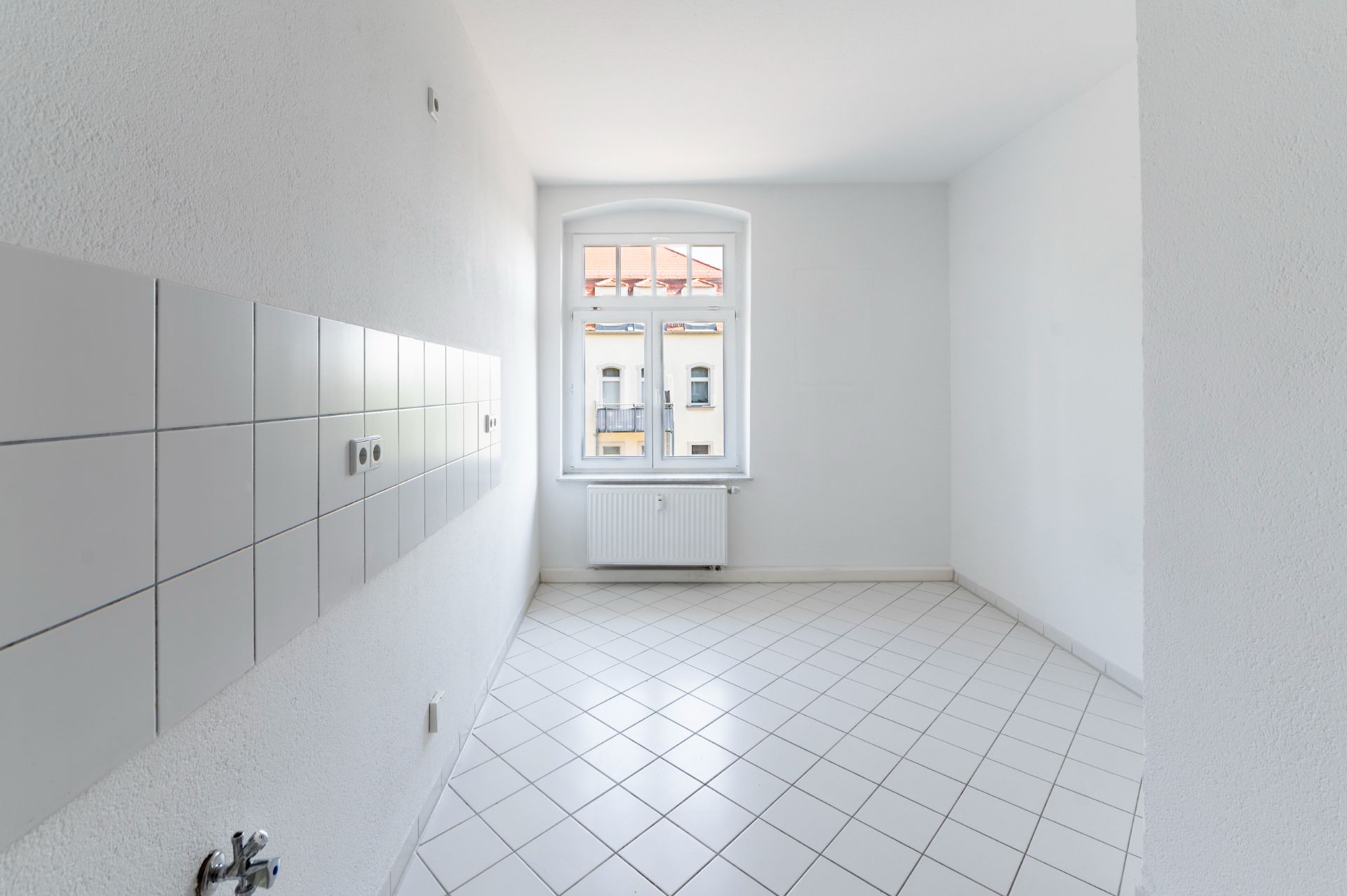 Heller, leerer Raum mit weißen Fliesen, Fenster und Heizkörper an der Wand.