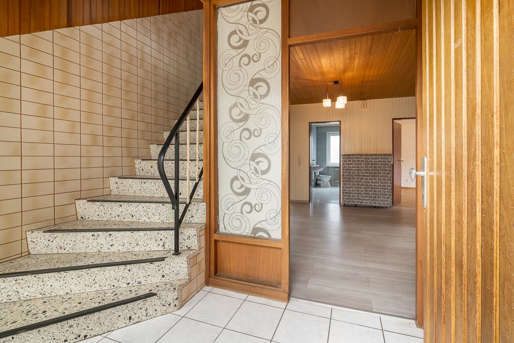 Eingangsbereich mit Treppe, Fliesenboden und Blick in ein Wohnzimmer mit Holzwand und Deckenleuchte.