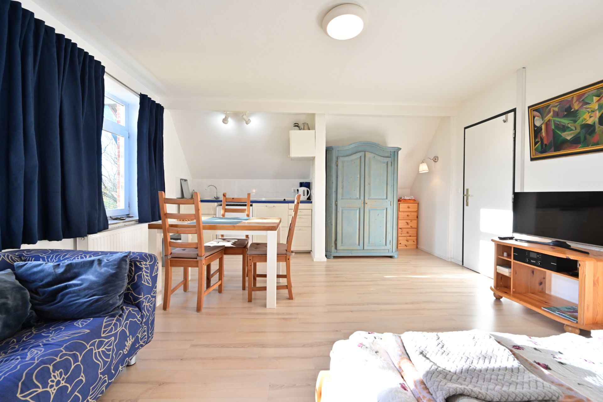 Helles Wohnzimmer mit Sofa, Esstisch, blauen Vorhängen, TV, Schrank und Küchenzeile.