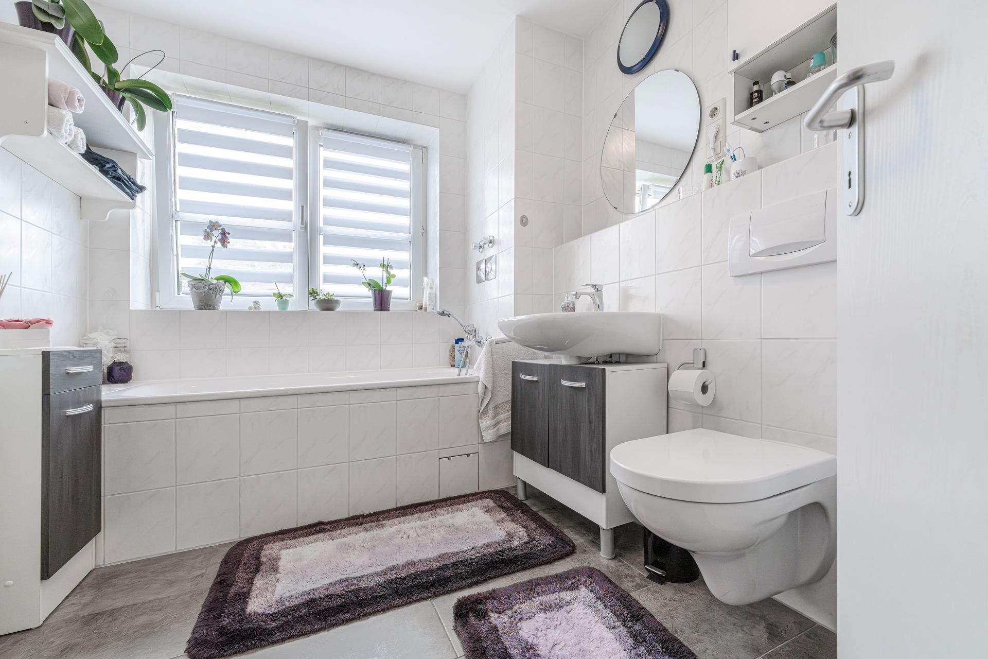 Helles Badezimmer mit Badewanne, Waschbecken, WC, zwei Fenstern und Teppichen auf dem Boden.