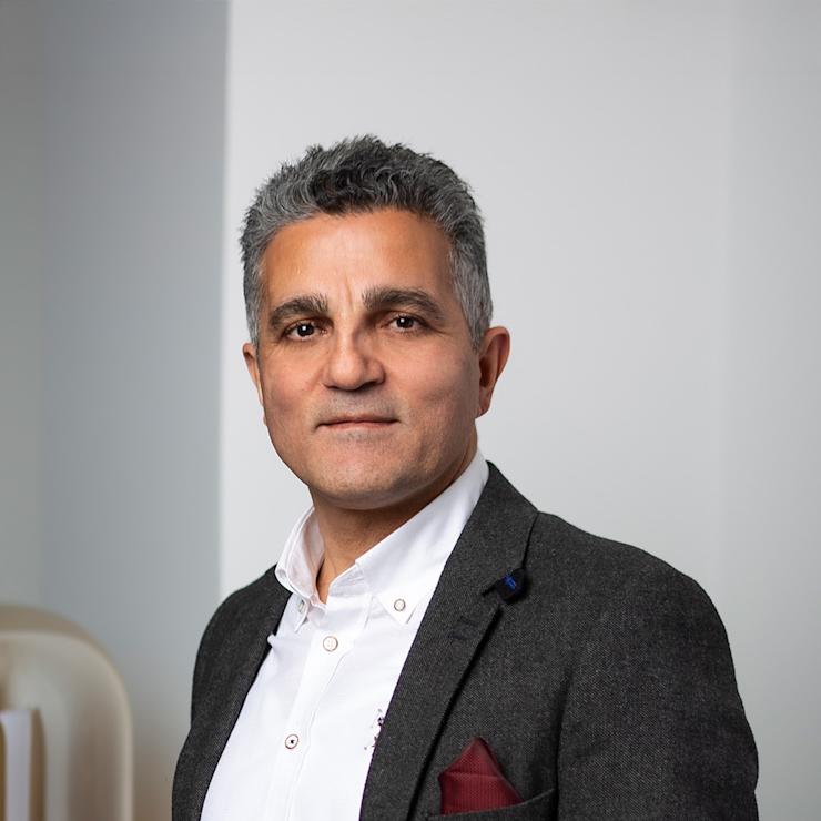 Ein Mann steht vor einer weißen Wand und trägt ein dunkles Sakko mit weißem Hemd.