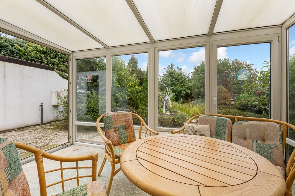 Wintergarten mit Holztisch und Stühlen, große Fenster mit Blick auf Garten.