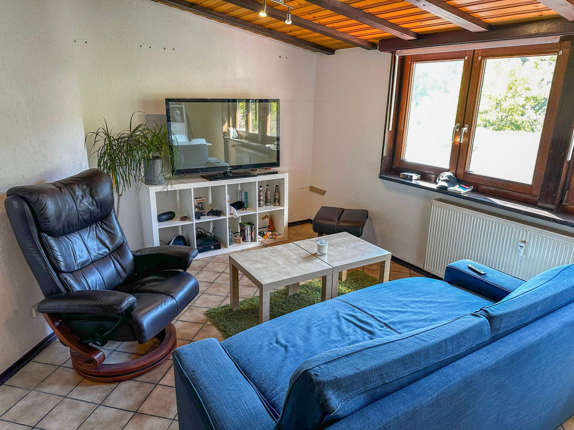 Wohnzimmer mit blauem Sofa, schwarzem Sessel, Couchtisch und Fernseher an der Fensterseite.