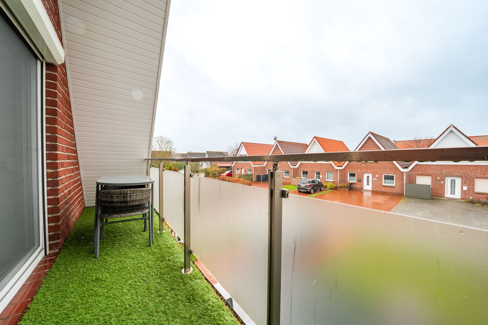 Schmaler Balkon mit Kunstrasen, Tisch und Stühlen, Blick auf Wohnhäuser mit roten Dächern.