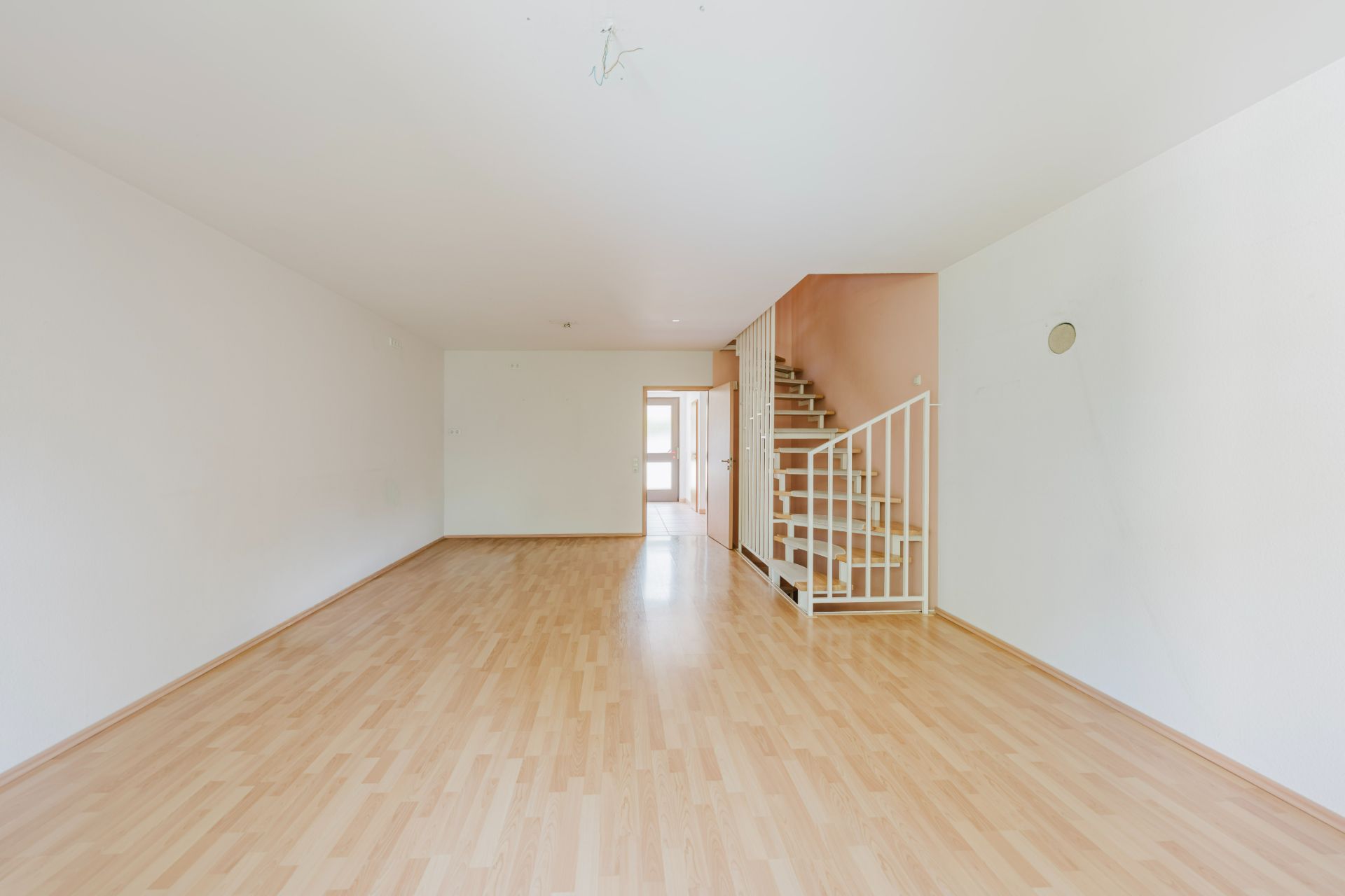 Leerer Raum mit hellem Holzfußboden und offener Treppe an der rechten Wand.