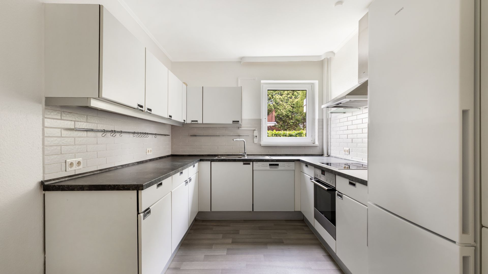 Moderne weiße Einbauküche mit Fenster, Elektrogeräten und hellgrauem Boden.