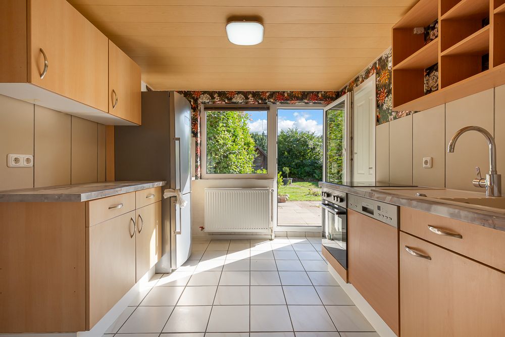 Helle Küche mit Holzmöbeln, Fliesenboden und Blick in einen grünen Garten durch offene Tür.