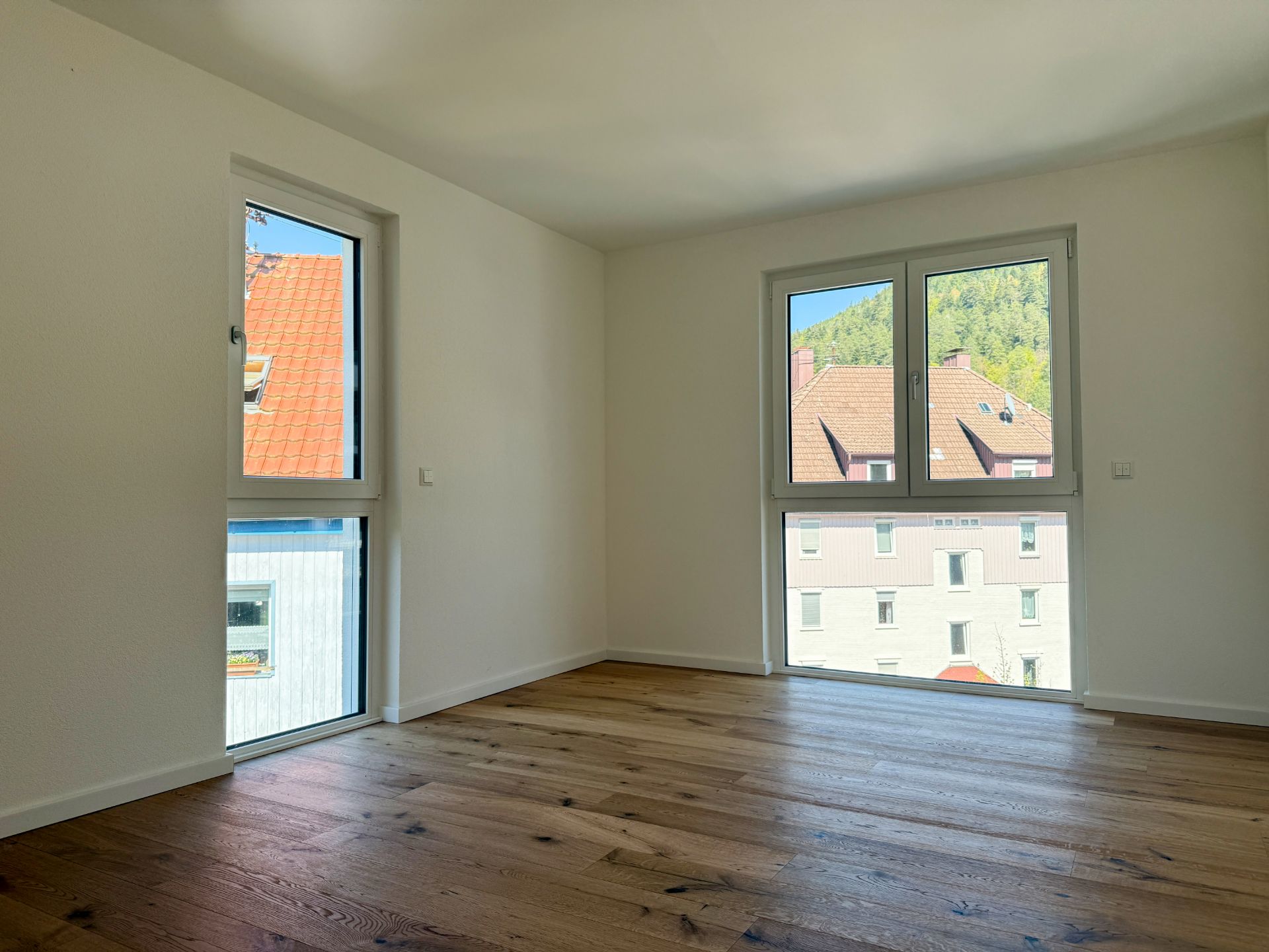 Helles Zimmer mit Holzfußboden und zwei großen Fenstern, Blick auf Nachbarhäuser.