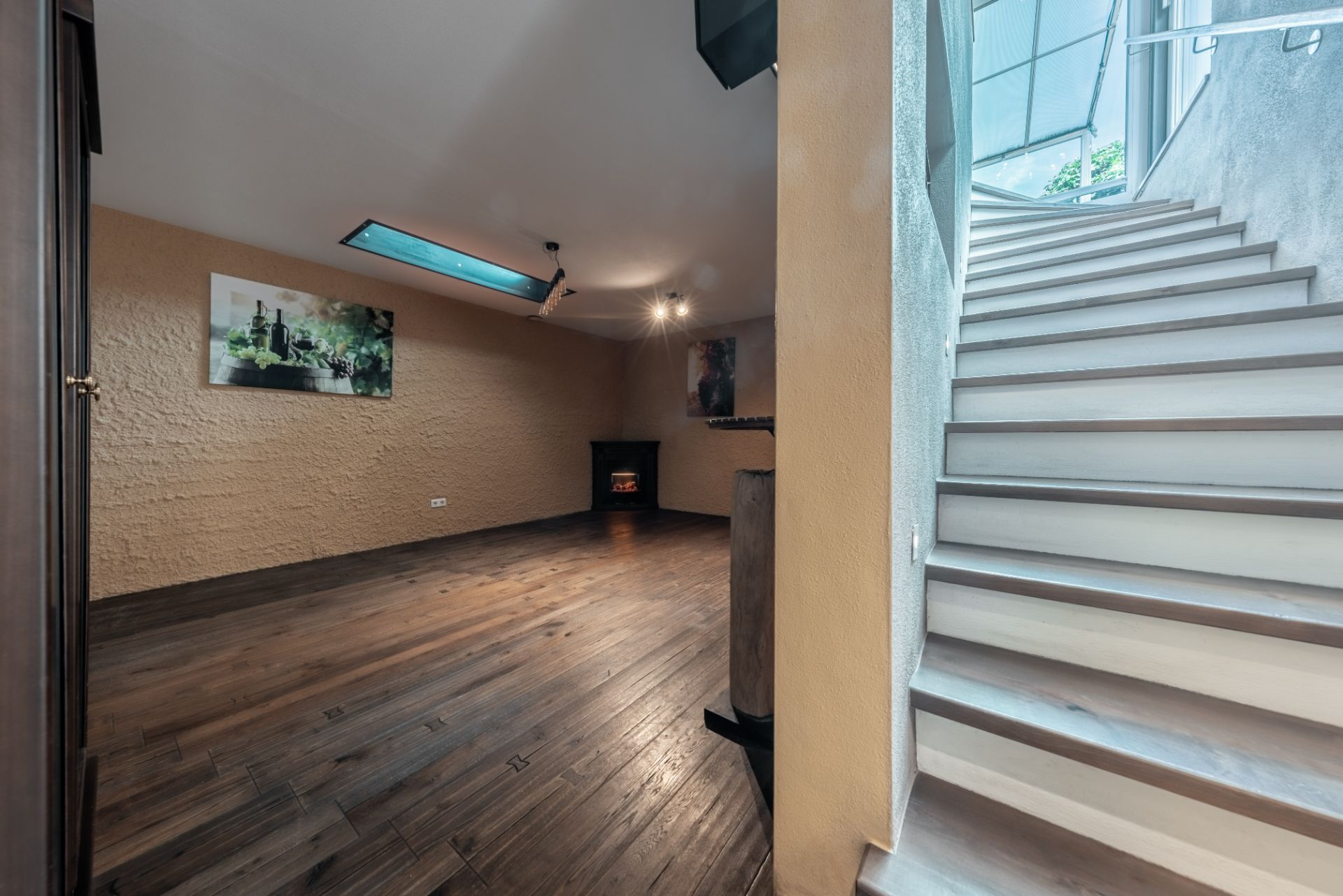 Raum mit Holzfußboden, Kamin und Bildern an der Wand, daneben eine Treppe mit Tageslicht.