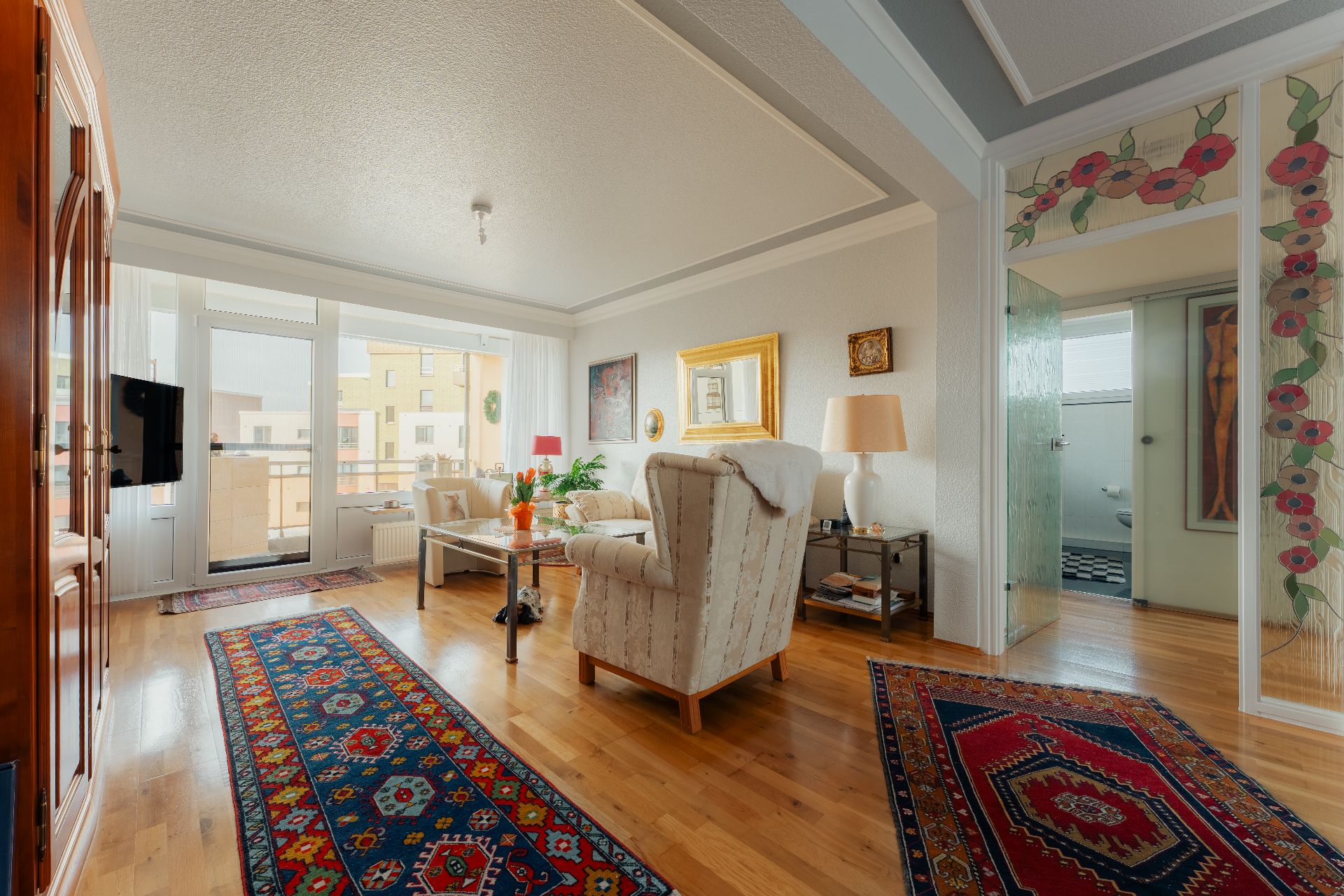 Helles Wohnzimmer mit Teppichen, Sesseln, Holzboden und Zugang zum Balkon.
