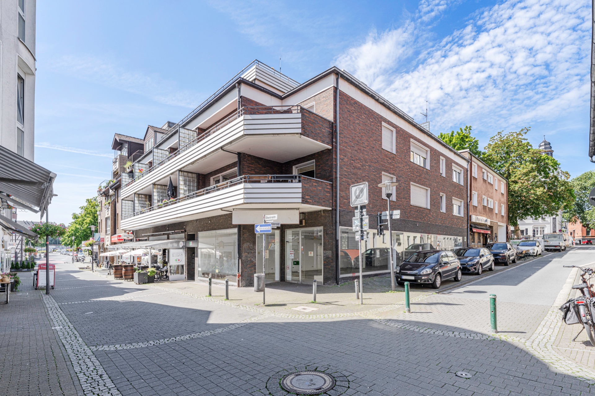 Eckwohnhaus mit Balkonen, Backsteinfassade und geparkten Autos an einer ruhigen Straße.