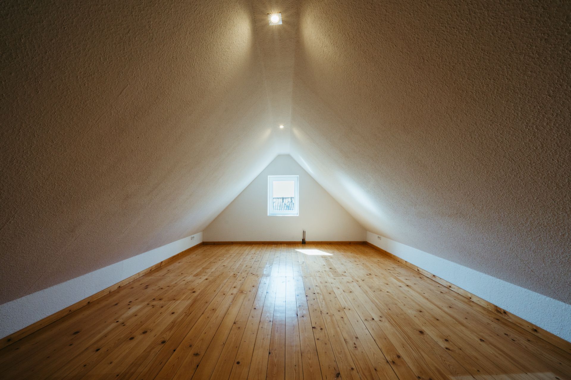 Dachgeschossraum mit Holzfußboden, schrägen Wänden und kleinem Fenster am Ende.