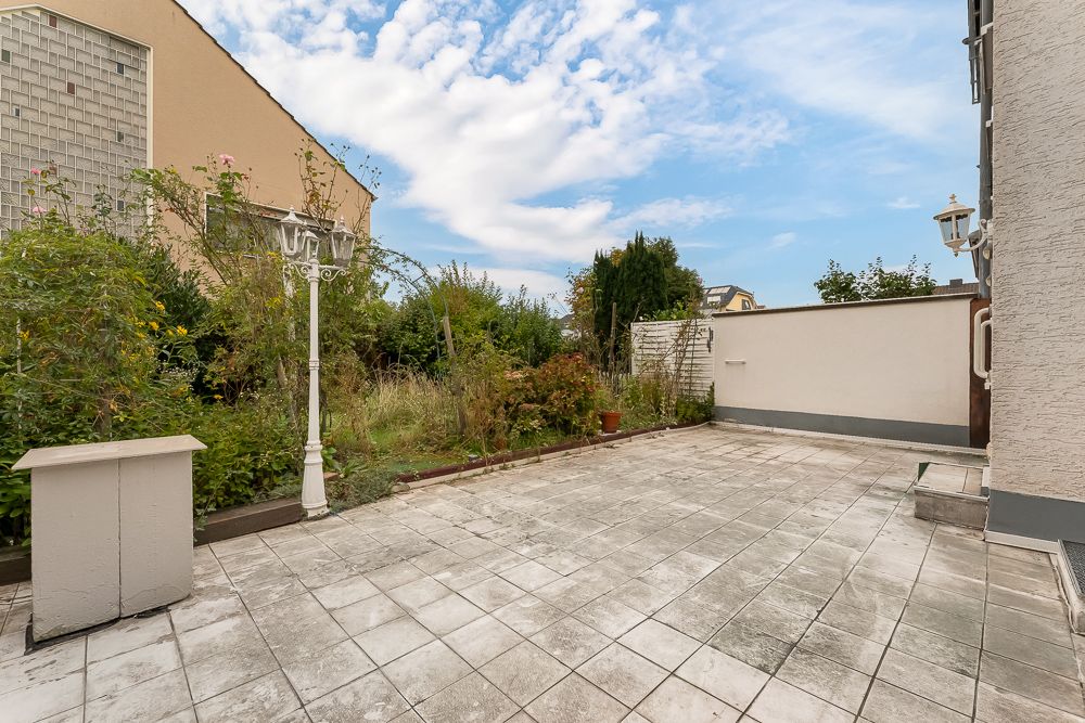 Geflieste Terrasse mit weißen Lampen, angrenzend an einen Garten mit Büschen und einer Mauer.