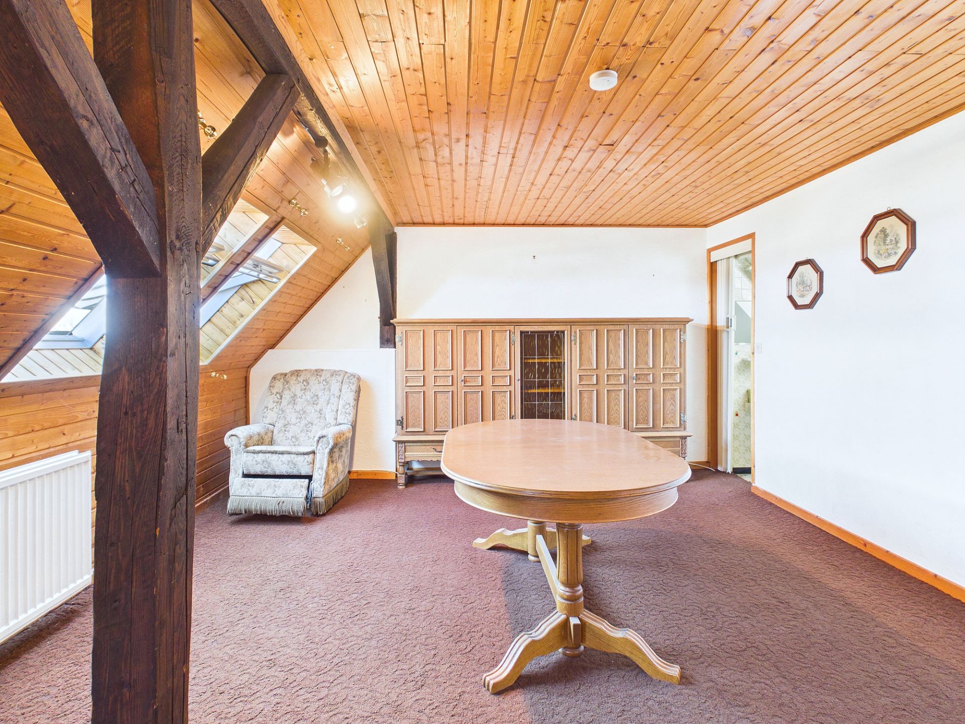 Wohnzimmer mit Holzdecke, rundem Tisch, Sessel und Holzschrank unter Dachschrägen.