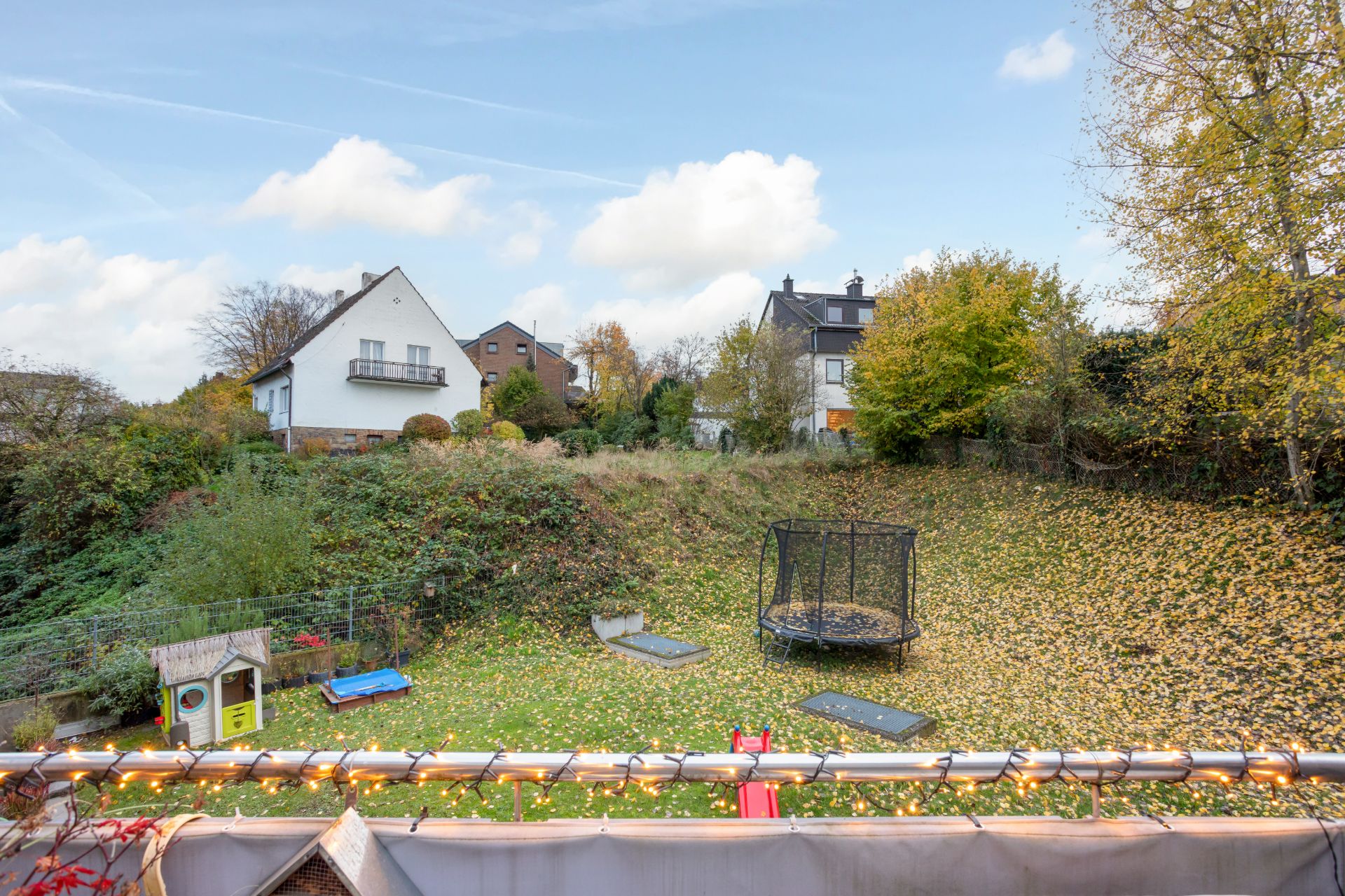 Garten mit Trampolin, Rutsche und Spielhaus, umgeben von Bäumen und Nachbarhäusern.