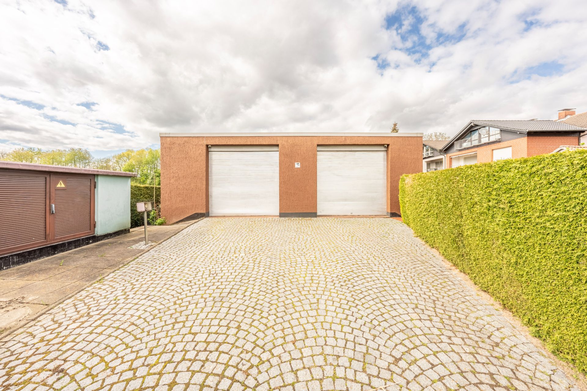 Doppelgarage mit zwei Rolltoren, gepflasterte Einfahrt und hohe Hecke rechts.