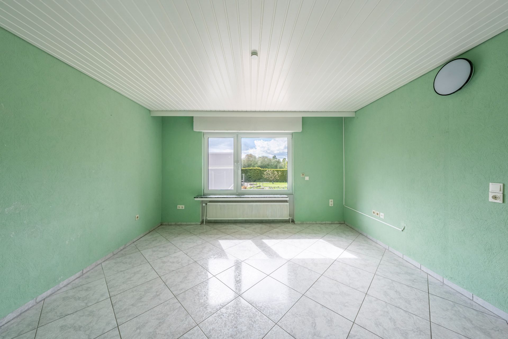 Leerer Raum mit grünen Wänden, weißer Decke, Fliesenboden und Fenster mit Blick ins Grüne.