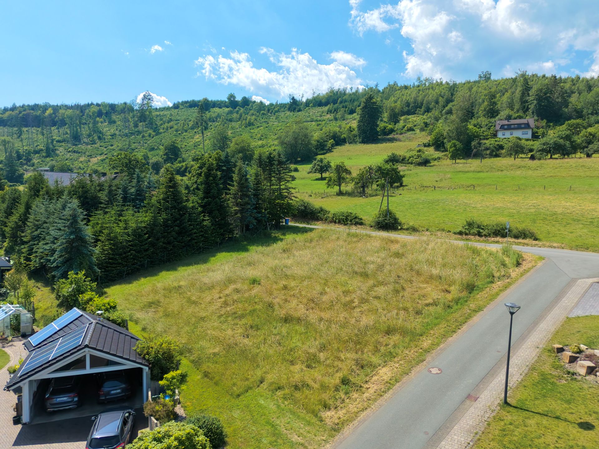 Leeres, grasbewachsenes Grundstück an einer ruhigen Straße mit Bäumen und Hügeln im Hintergrund.