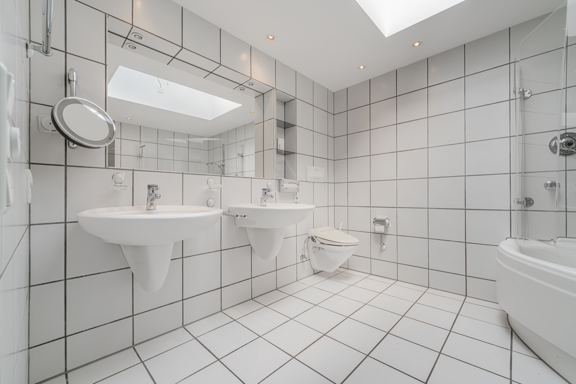 Modernes Badezimmer mit zwei Waschbecken, einem WC und einer Badewanne, alles in weiß gefliest.