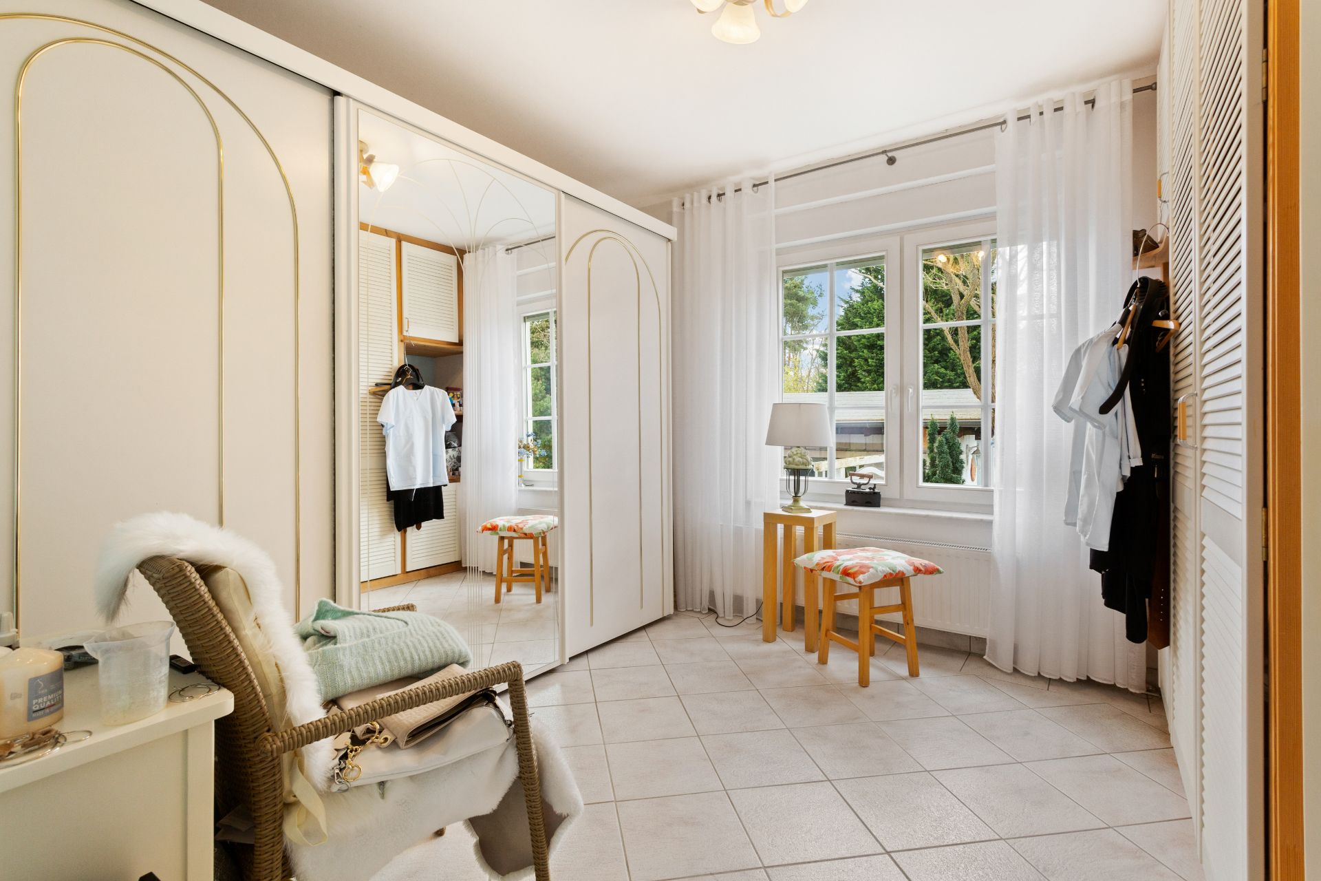 Heller Ankleideraum mit großem Fenster, Spiegel, Stuhl, Hockern und Garderobe.