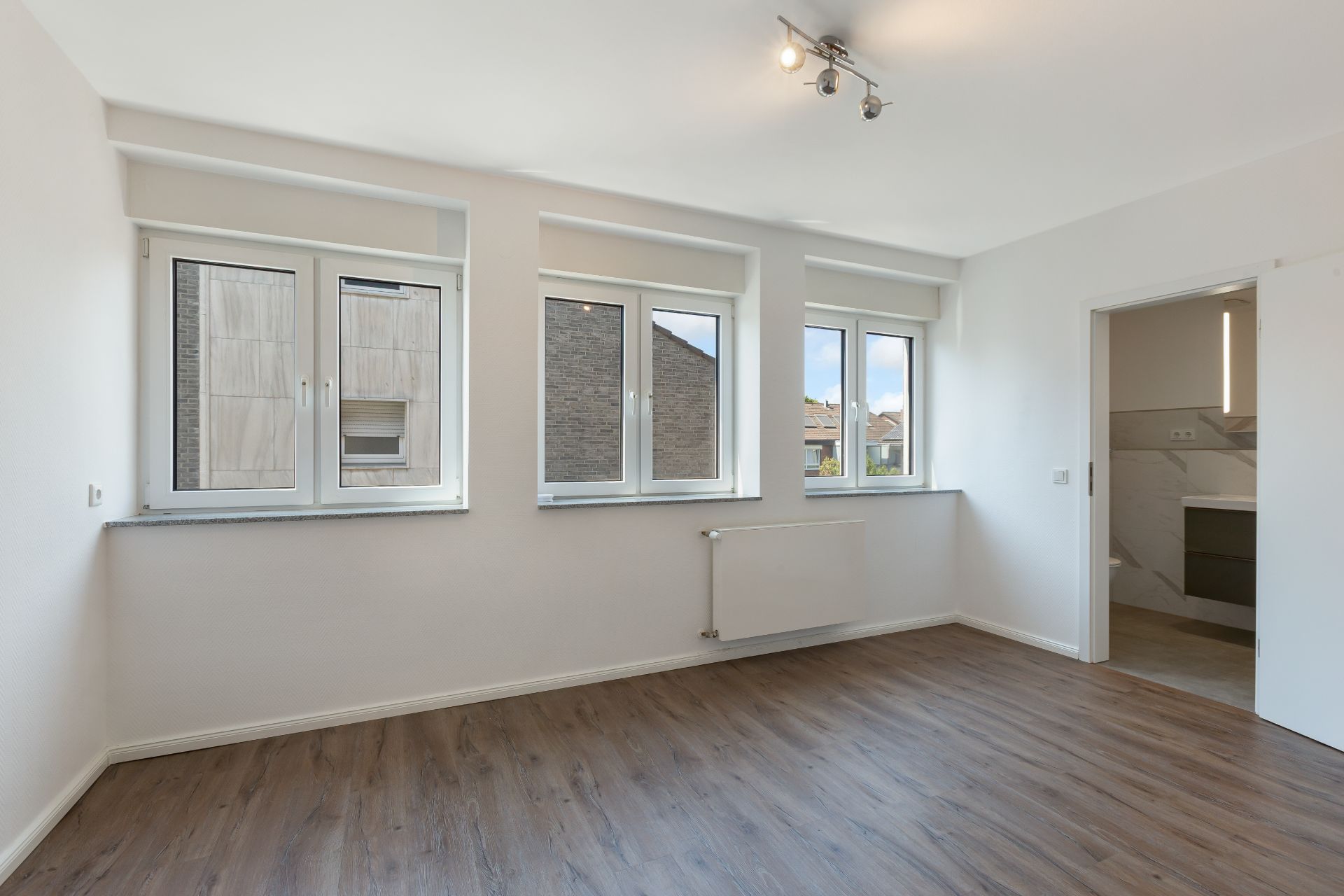 Leerer Raum mit drei Fenstern, Holzfußboden und Zugang zu einem Badezimmer.