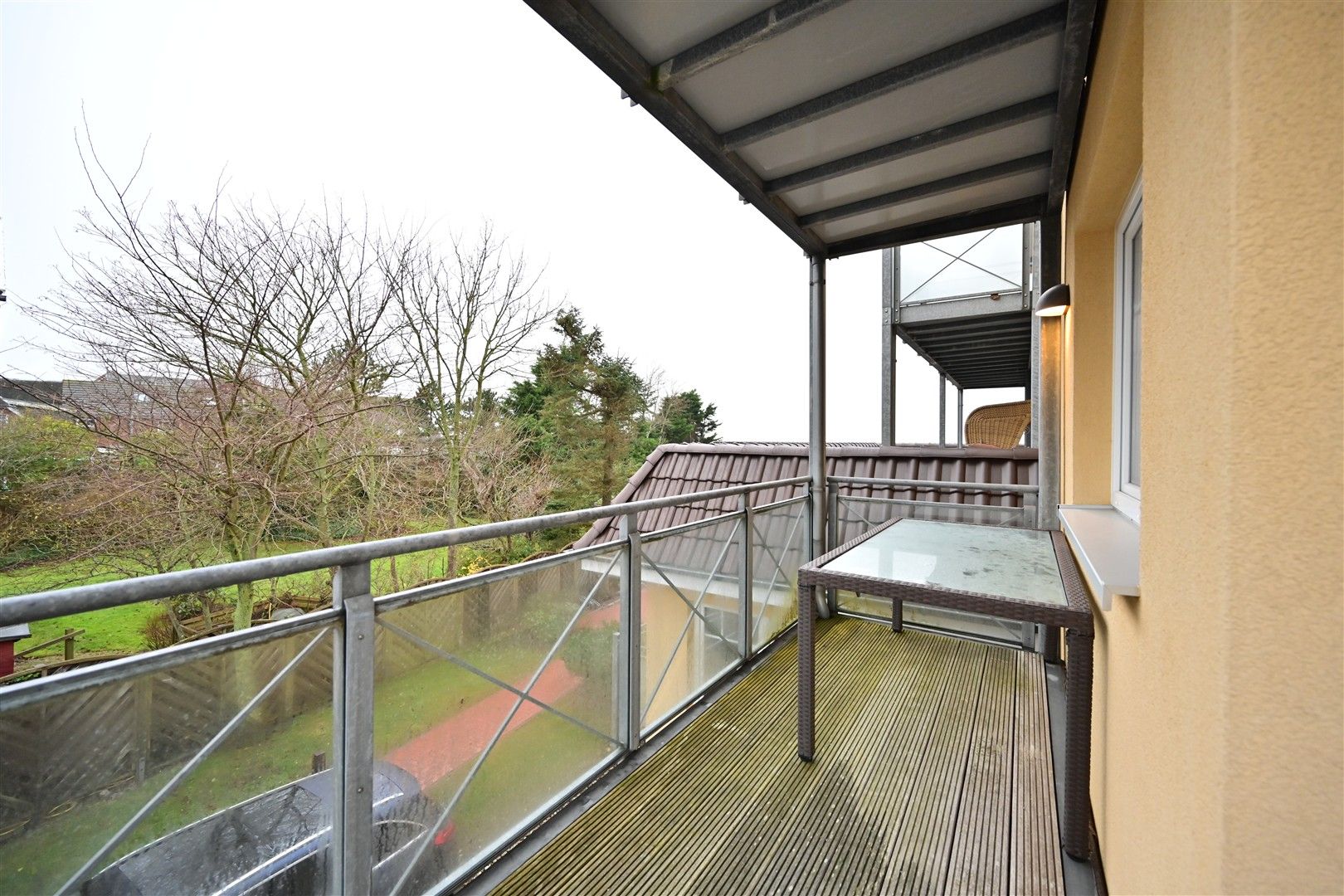 Überdachter Balkon mit Glasgeländer und Tisch, Blick auf Garten mit Bäumen.
