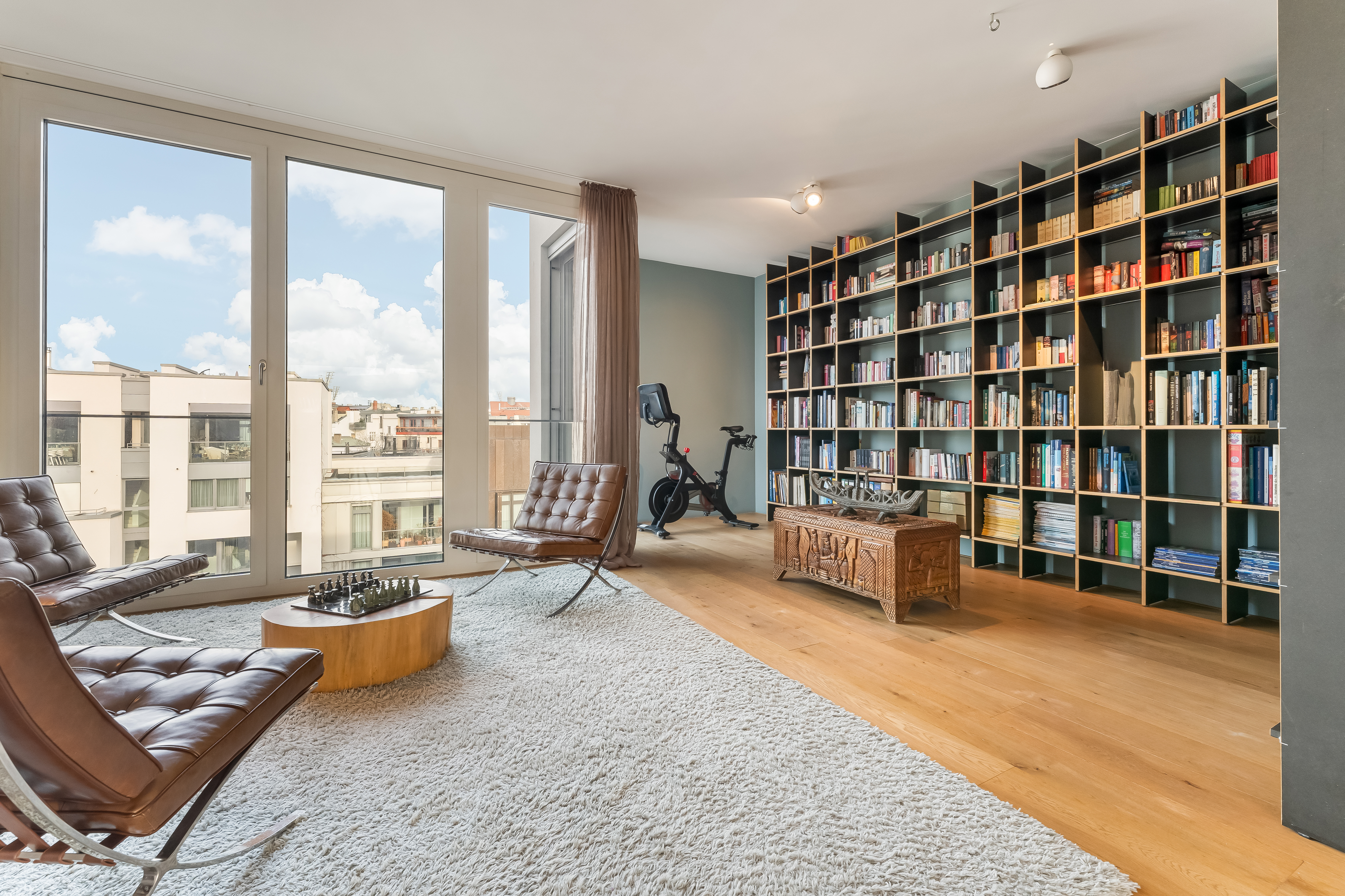 Helles Wohnzimmer mit Bücherregal, Sesseln, Teppich und großen Fenstern.