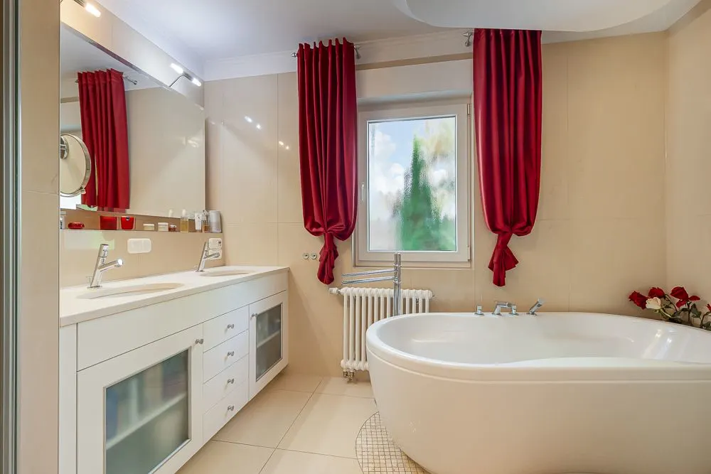 Modernes Badezimmer mit ovaler Badewanne, Doppelwaschbecken und roten Vorhängen am Fenster.