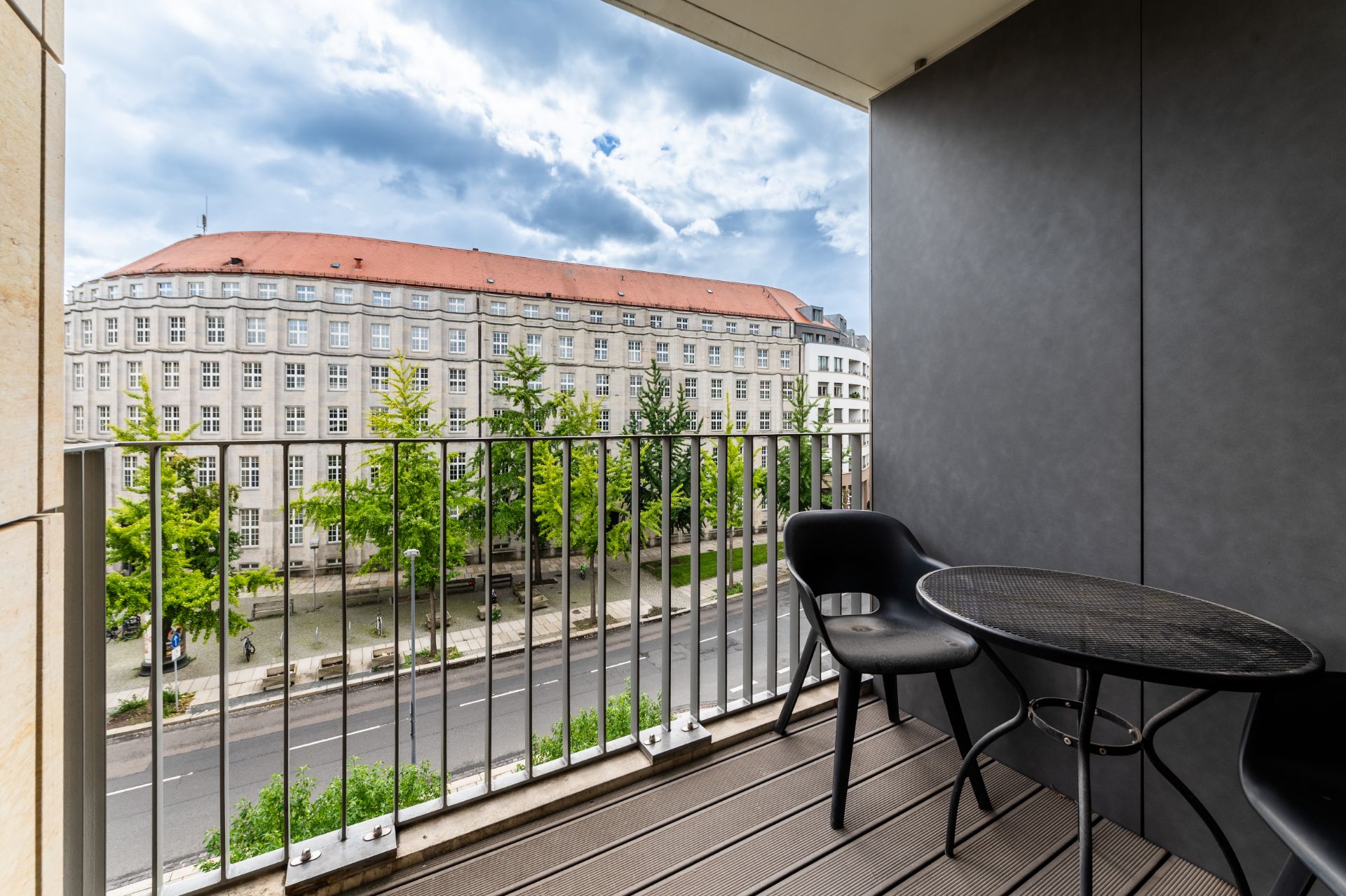 Balkon mit zwei schwarzen Stühlen und rundem Tisch, Blick auf eine Straße und ein großes Wohngebäude.