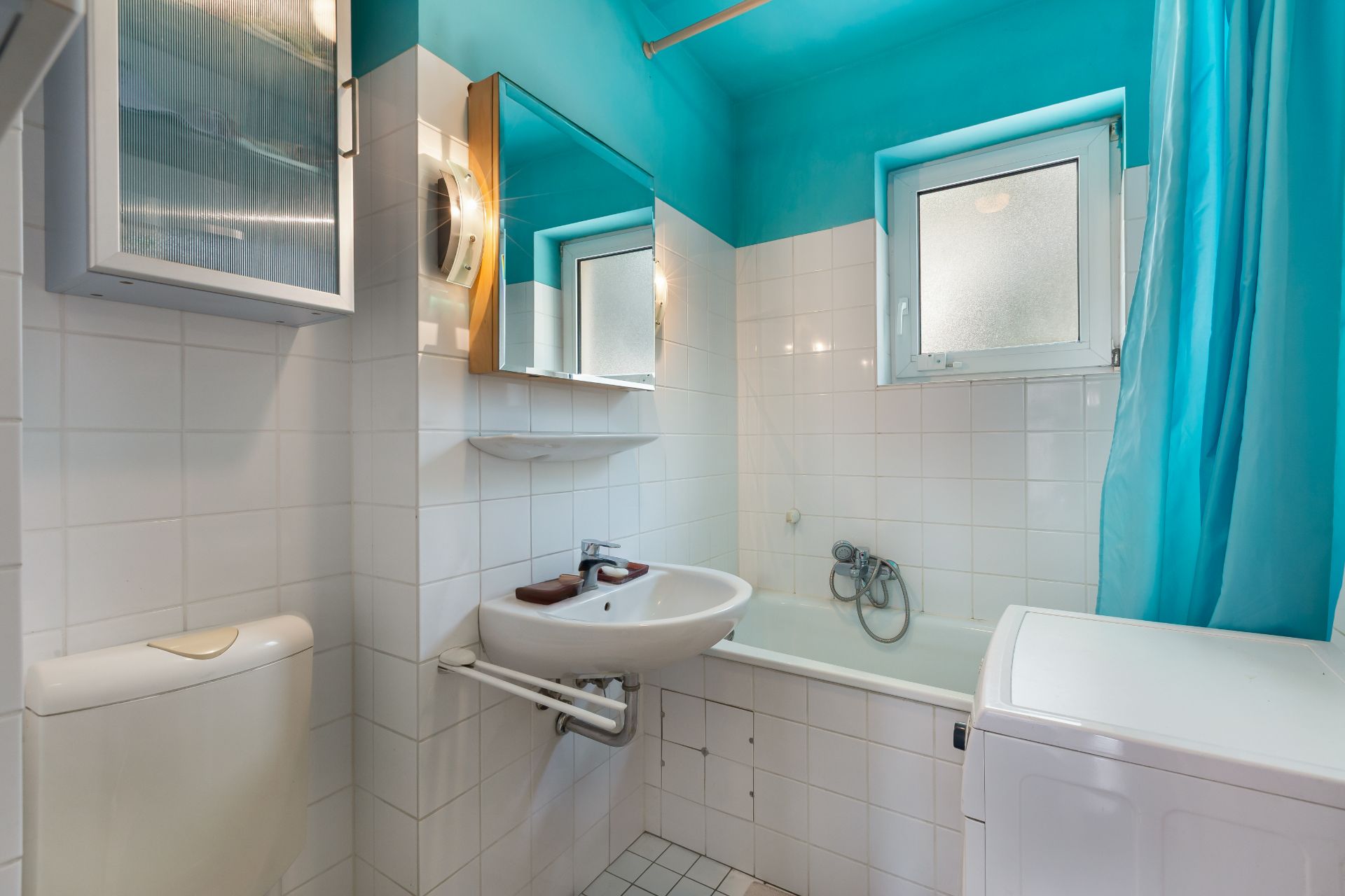 Kleines Badezimmer mit Waschbecken, Toilette, Badewanne, Waschmaschine und Fenster.