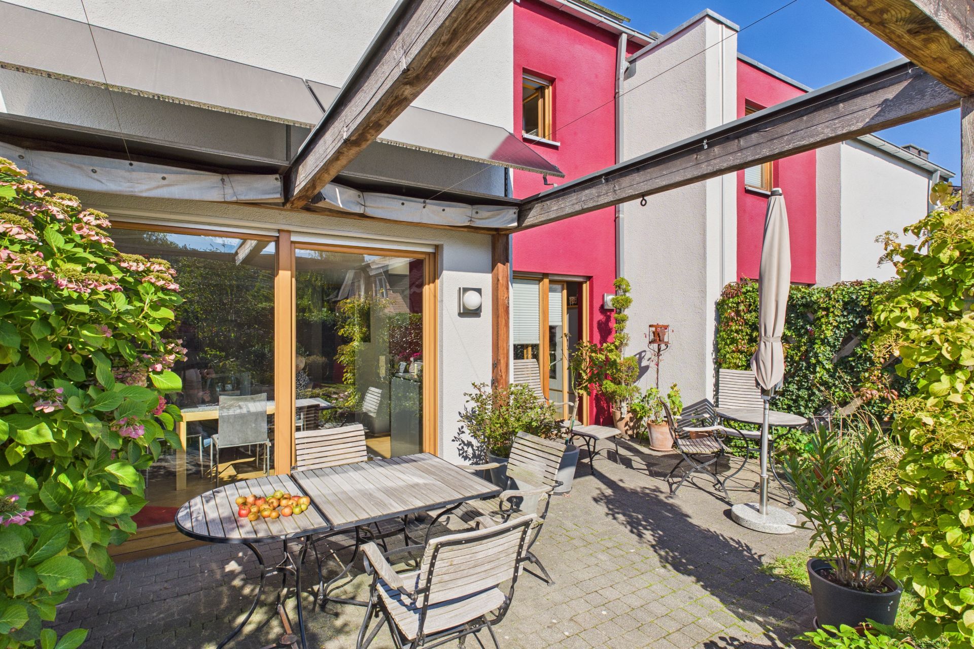Terrasse mit Tisch, Stühlen, Sonnenschirm und Pflanzen vor einem modernen Wohnhaus.