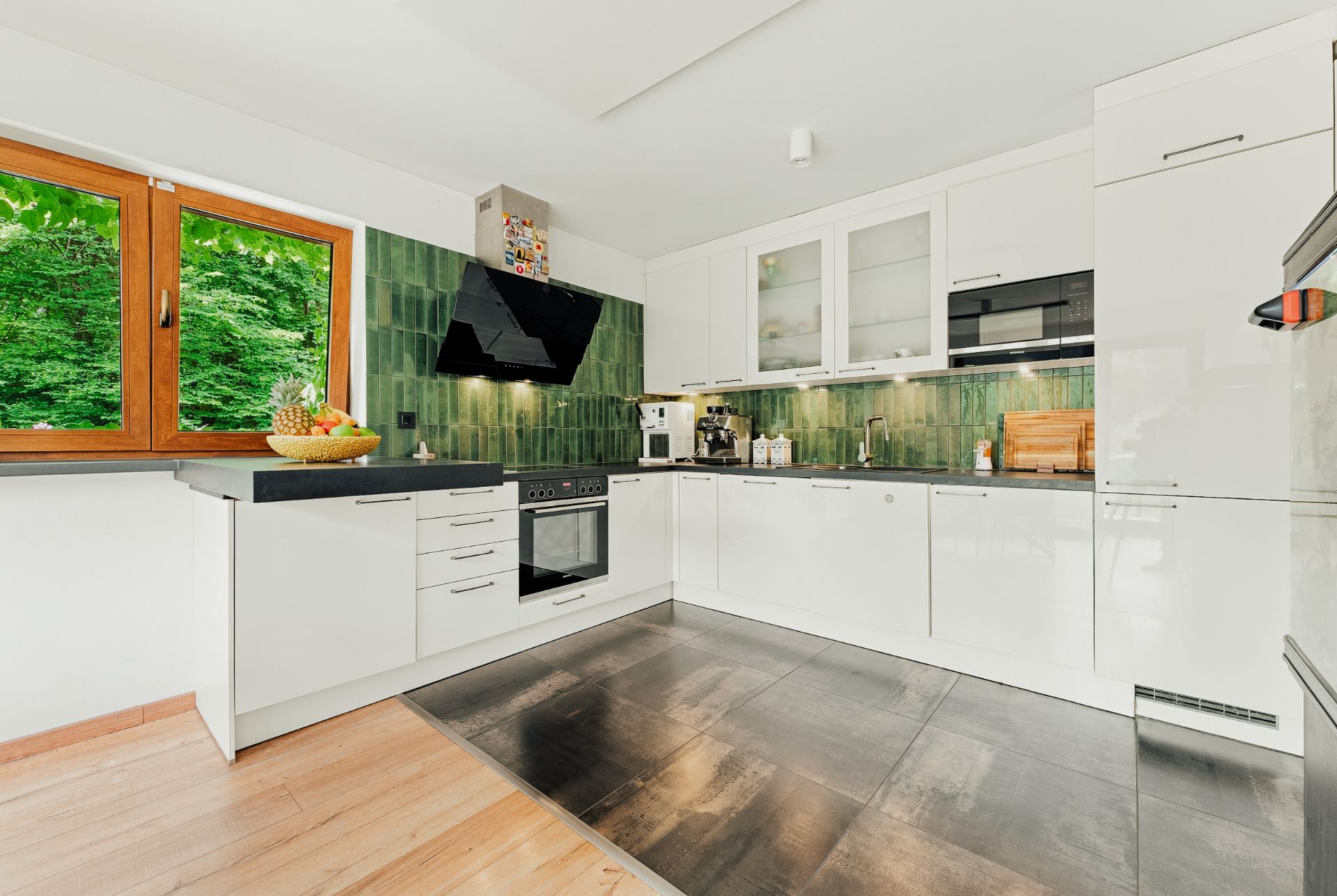 Moderne weiße Einbauküche mit grünem Fliesenspiegel und großem Fenster mit Blick ins Grüne.