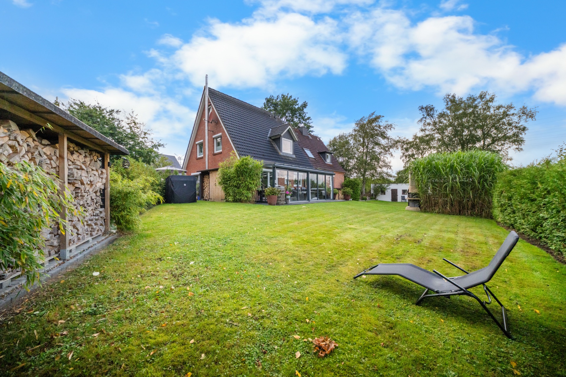 Haus mit Spitzdach, großer Garten, Liegestuhl und Holzunterstand links.