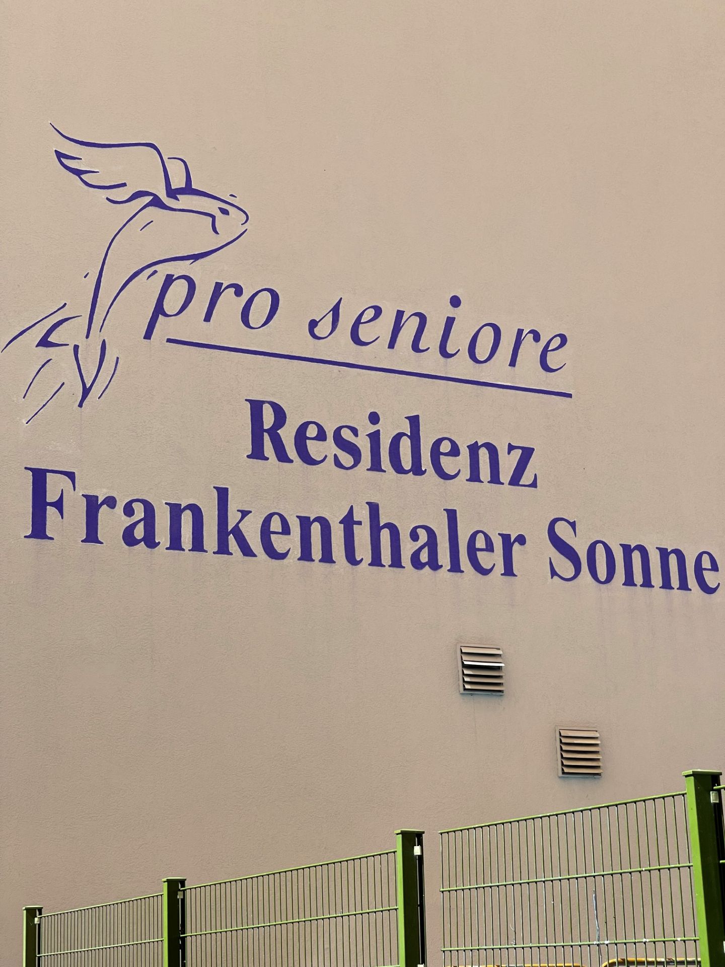 Gebäudewand mit lila Schriftzug 'pro seniore Residenz Frankenthaler Sonne' und Zaun im Vordergrund.
