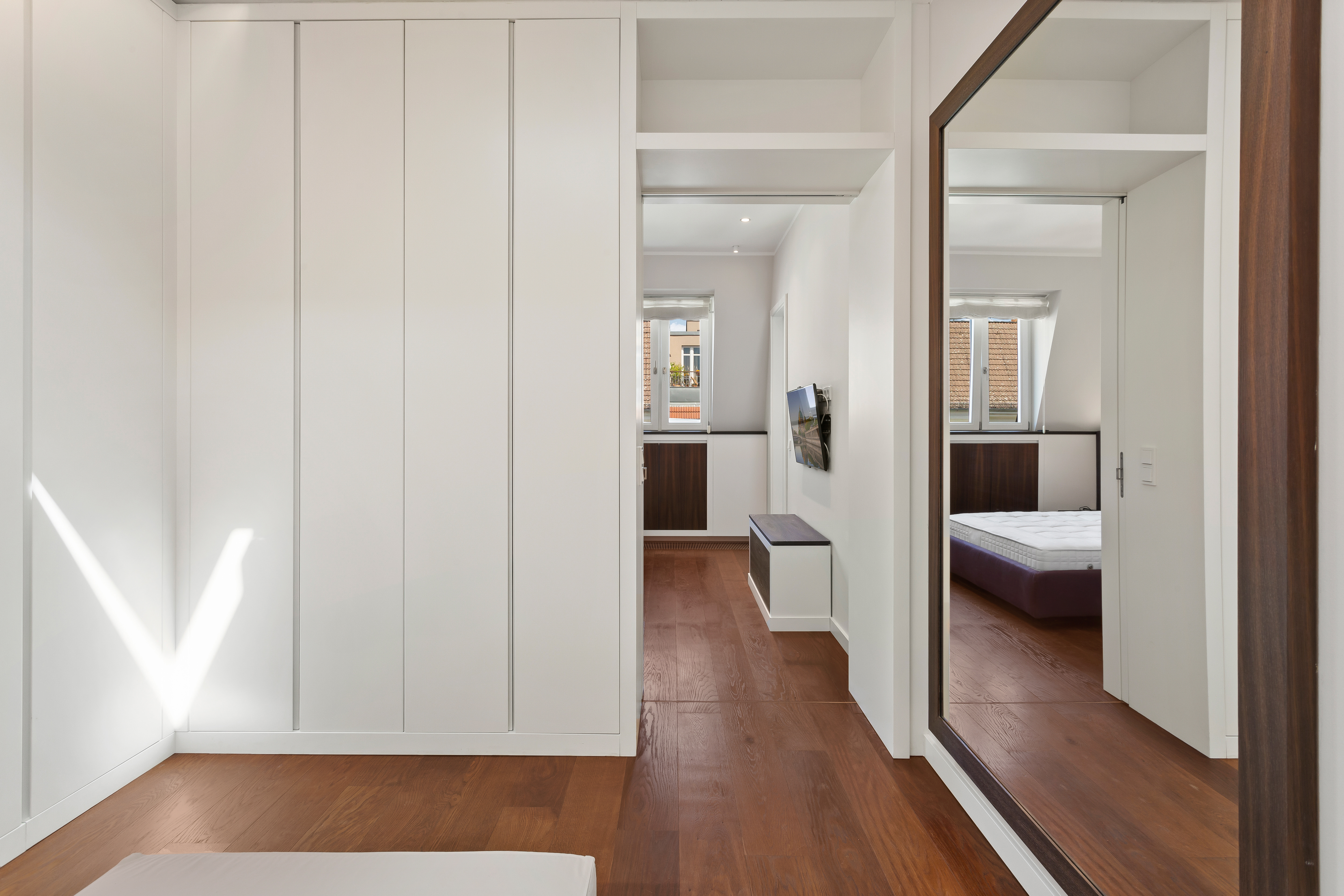 Heller Raum mit Holzfußboden, großem Spiegel, Einbauschrank und Blick auf ein Bett im Nebenraum.