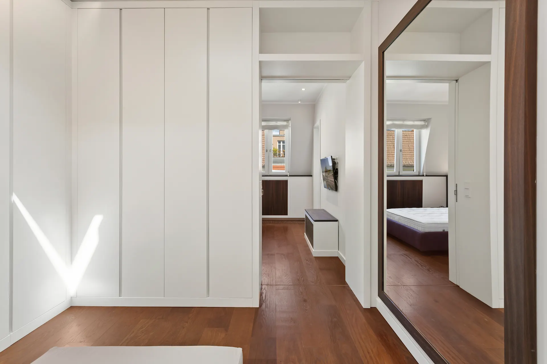 Heller Raum mit Holzfußboden, großem Spiegel, Einbauschrank und Blick auf ein Bett im Nebenraum.