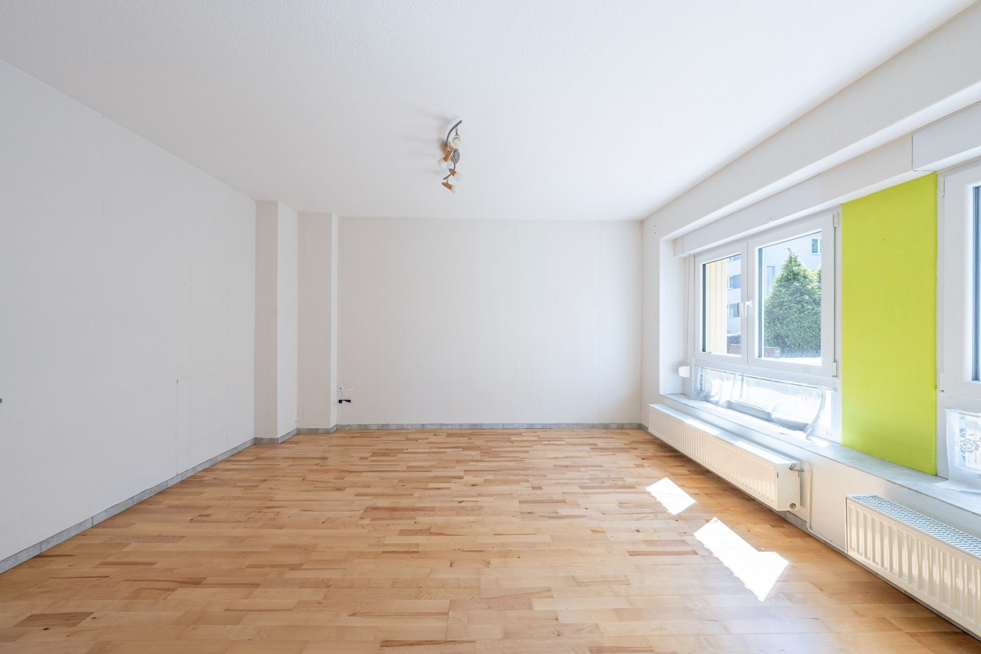 Helles, leeres Zimmer mit Holzboden und großen Fenstern, ein grünes Fensterpanel sichtbar.