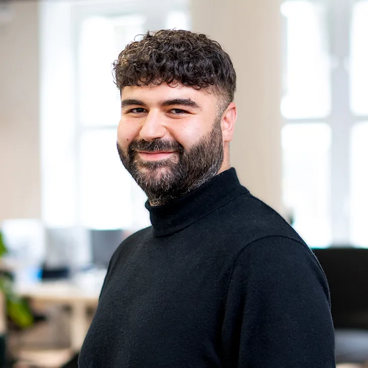 Ein Mann mit Bart und dunklem Pullover steht lächelnd in einem hellen Raum mit großen Fenstern.