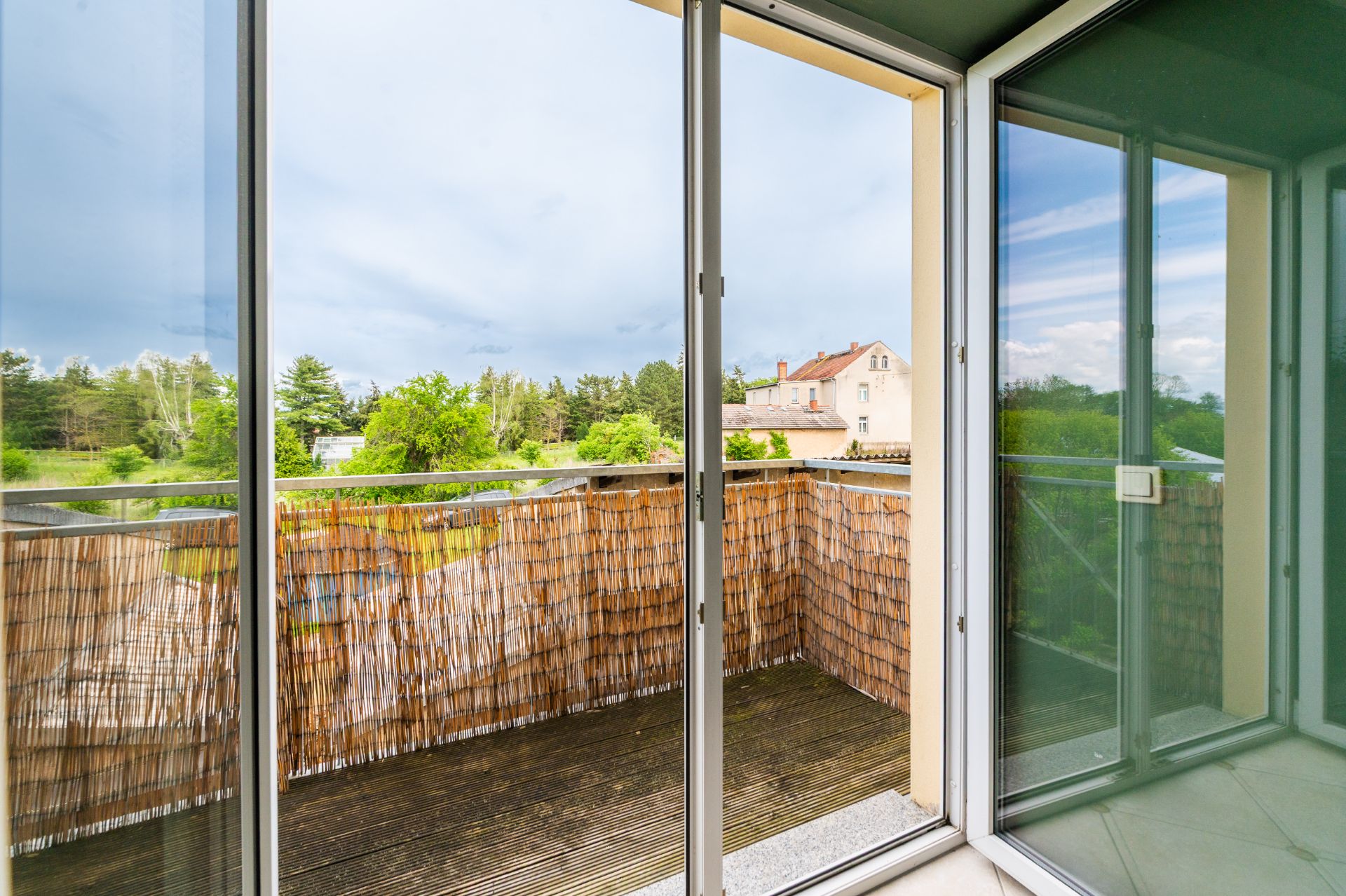 Balkon mit Holzsichtschutz und Ausblick auf Bäume und ein Wohnhaus, fotografiert durch eine Glas-Schiebetür.