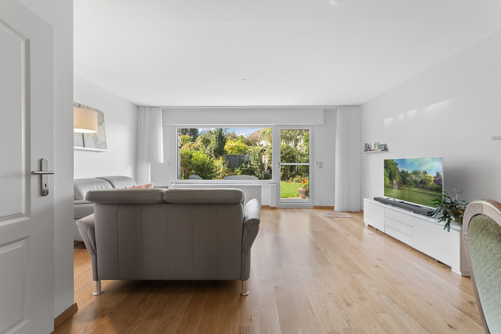 Helles Wohnzimmer mit grauem Sofa, Fernseher und Blick auf einen Garten durch große Fenster.