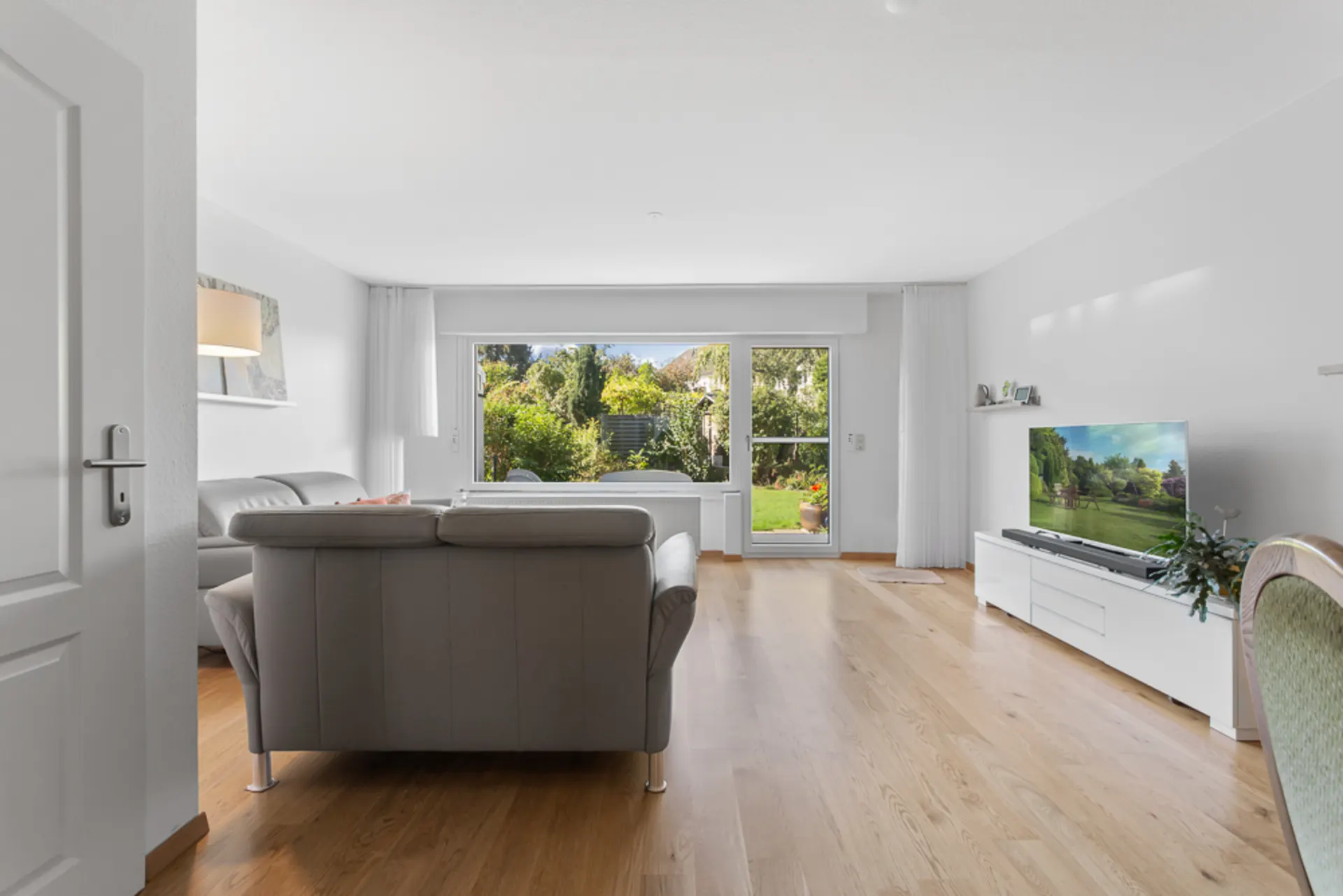 Helles Wohnzimmer mit grauem Sofa, Fernseher und Blick auf einen Garten durch große Fenster.