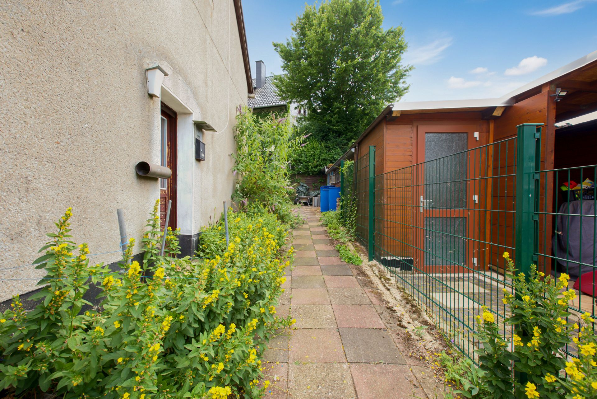 Gepflasterter Weg mit gelben Blumen, links Hauswand, rechts Gartenhaus und Zaun.