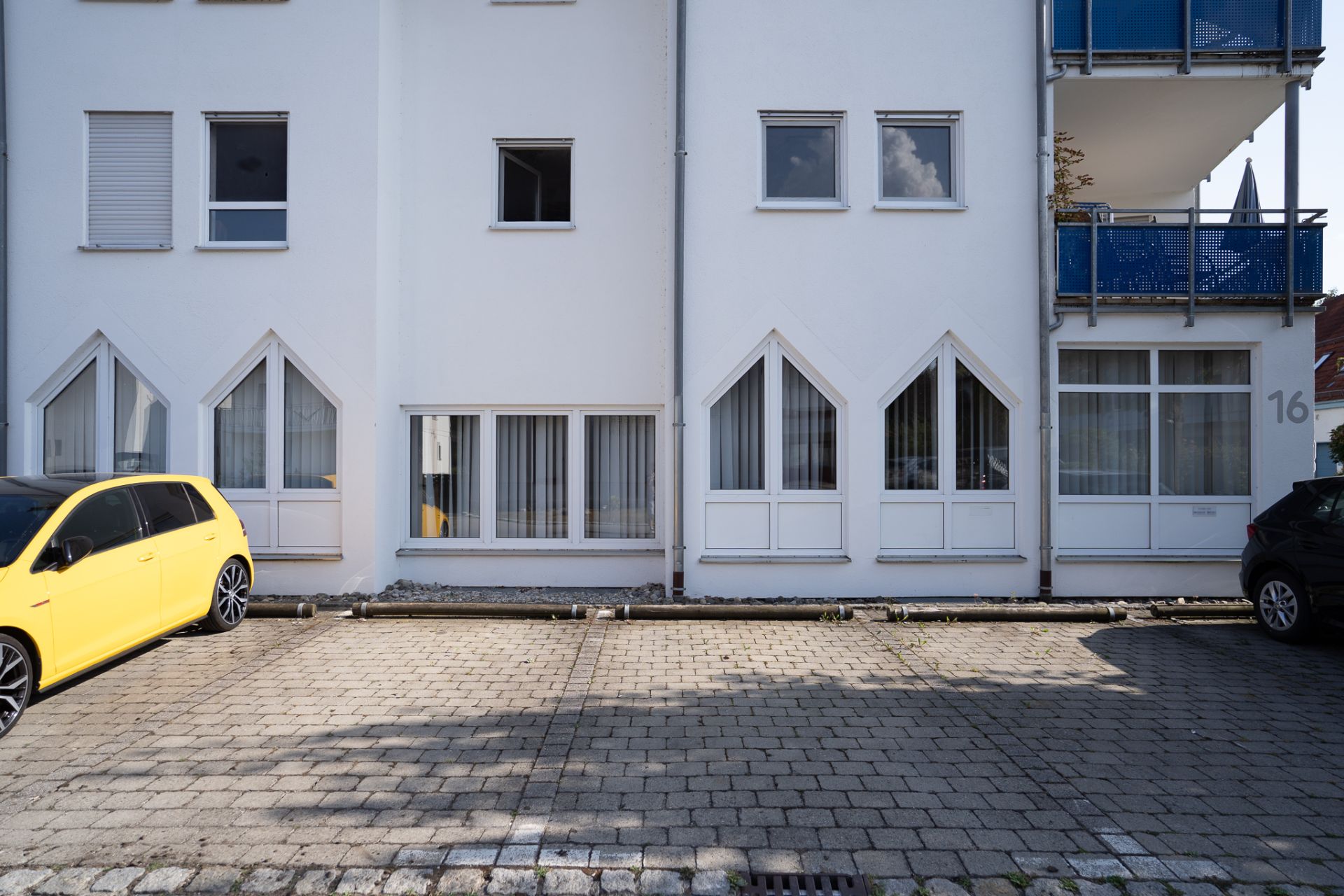 Weiße Wohnhausfassade mit mehreren Fenstern und geparkten Autos auf gepflastertem Parkplatz.