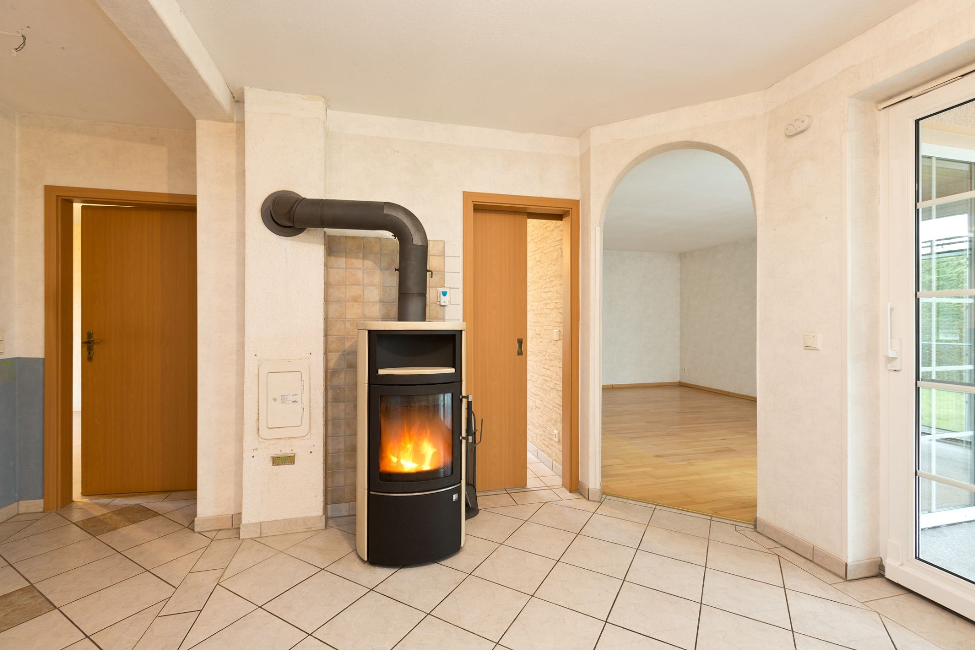 Raum mit Kaminofen auf Fliesenboden, zwei Holztüren und Durchgang zum Wohnzimmer mit Parkett.