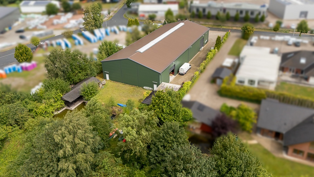 Luftaufnahme einer großen grünen Halle mit Gartenflächen und Bäumen rundherum.