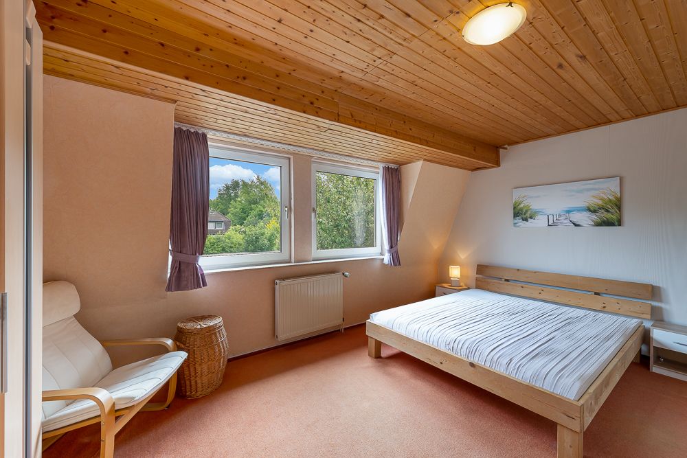 Helles Schlafzimmer mit Holzdecke, Doppelbett, Sessel, zwei Fenstern und Nachttischlampe.