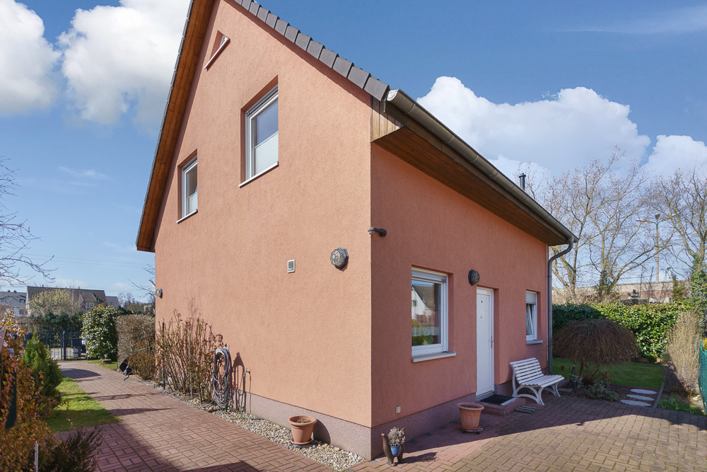 Einfamilienhaus mit rosafarbener Fassade, weißen Fenstern und kleinem Vorgarten bei Tageslicht.