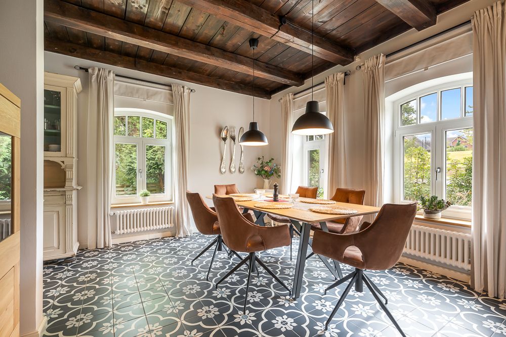 Esszimmer mit Holztisch, sechs Stühlen, großen Fenstern und Zierfliesenboden.