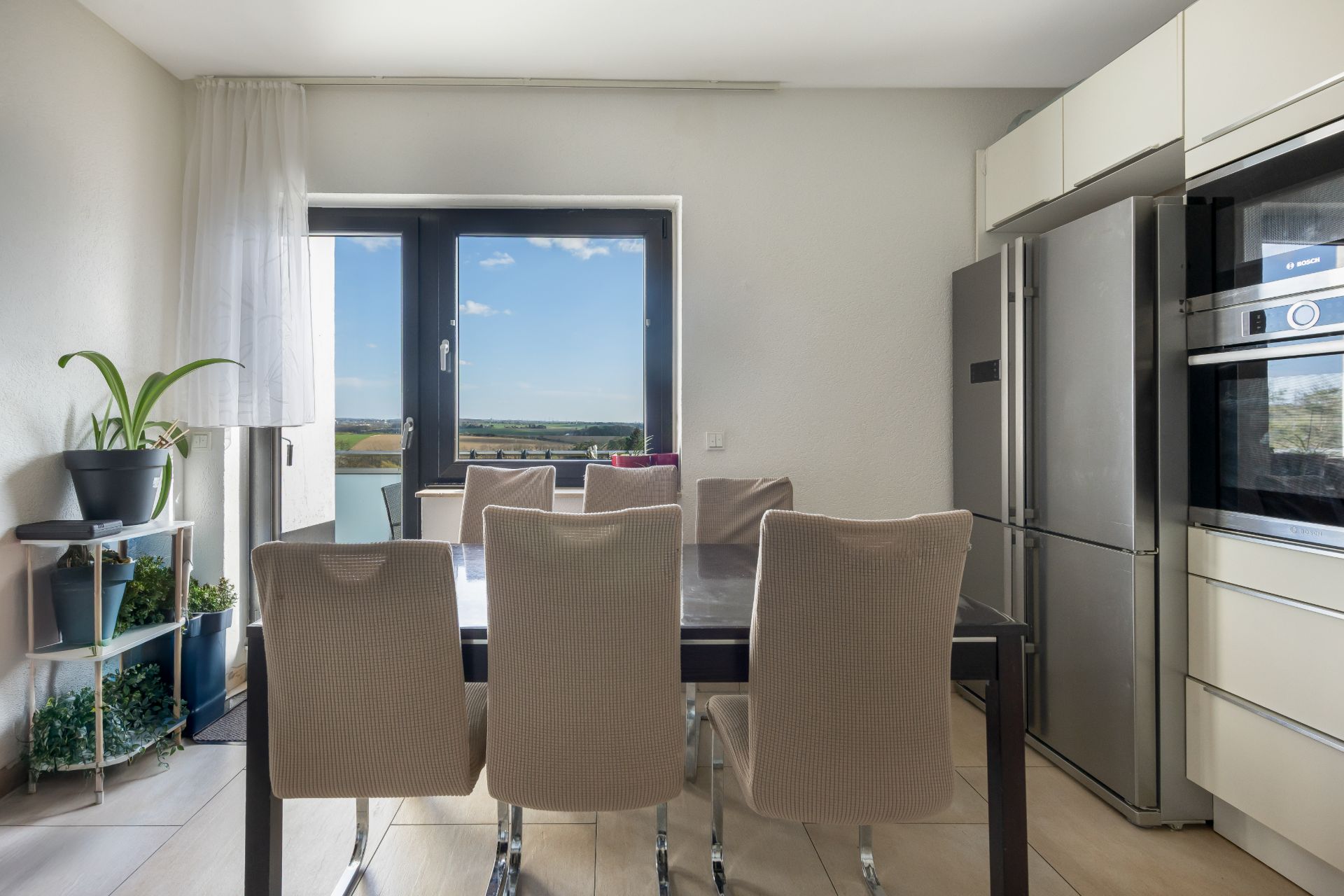 Esszimmer mit sechs Stühlen, großem Fenster, Pflanzenregal und moderner Küche.
